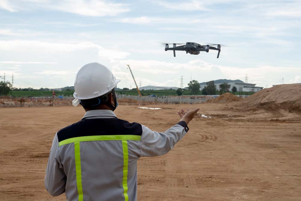 A construction worker in a white helmet and reflective jacket operates a drone at a construction site.