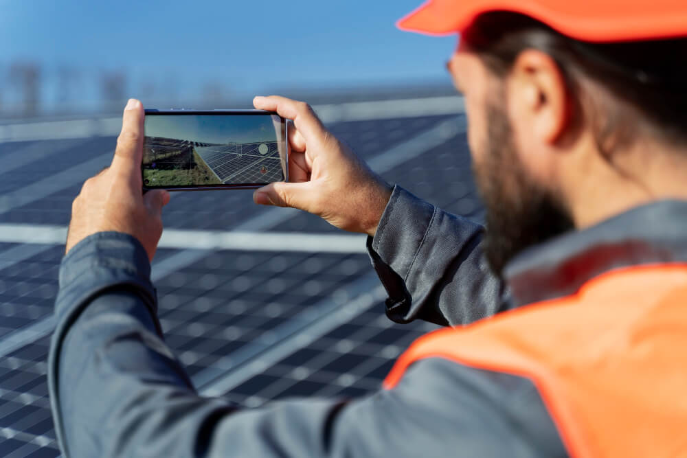 A worker in an orange vest and cap photographs solar panels with a smartphone. The scene conveys a focus on renewable energy and innovation.