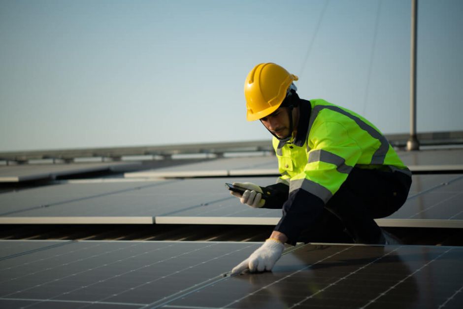 A worker in a yellow hard hat and reflective jacket inspects solar panels on a roof, using a smartphone. The scene conveys focus and modern technology.