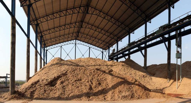 A large pile of sand is situated in the center of a spacious warehouse, surrounded by concrete walls and industrial lighting.
