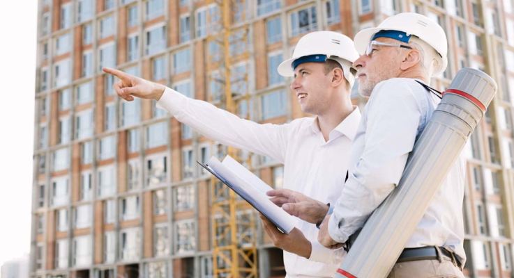 Two men in hard hats are pointing at an object, discussing details on a construction site.